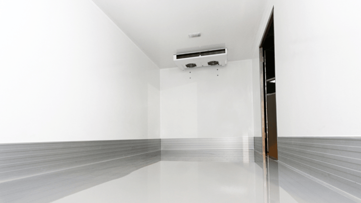 Inside of walk-in cooler - gray floor, silver flashing at bottom of walls, white walls and refrigeration unit on top of ceiling at far end